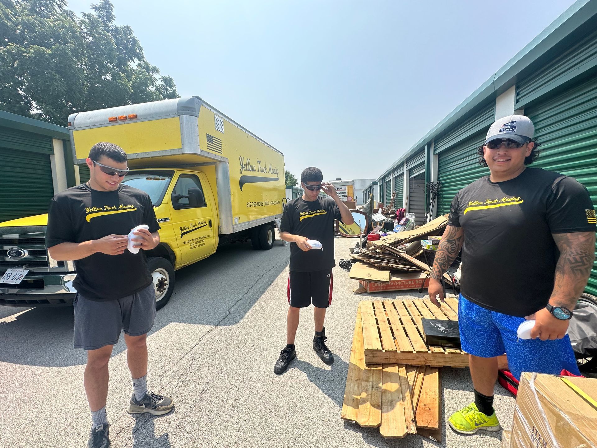 Three men near a yellow moving truck and storage units, holding wipes in the sun.