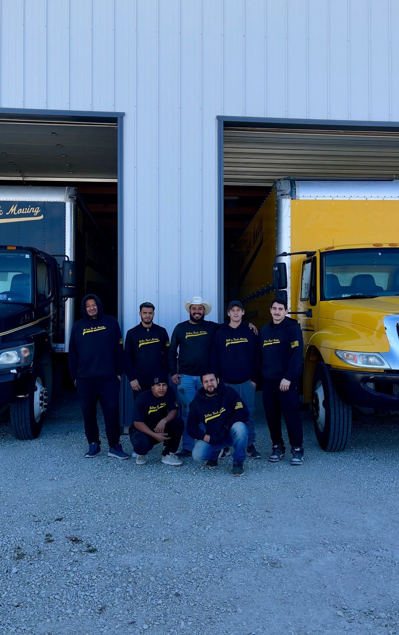 Team of movers posing in front of two trucks. They wear black hoodies, a building in the background.