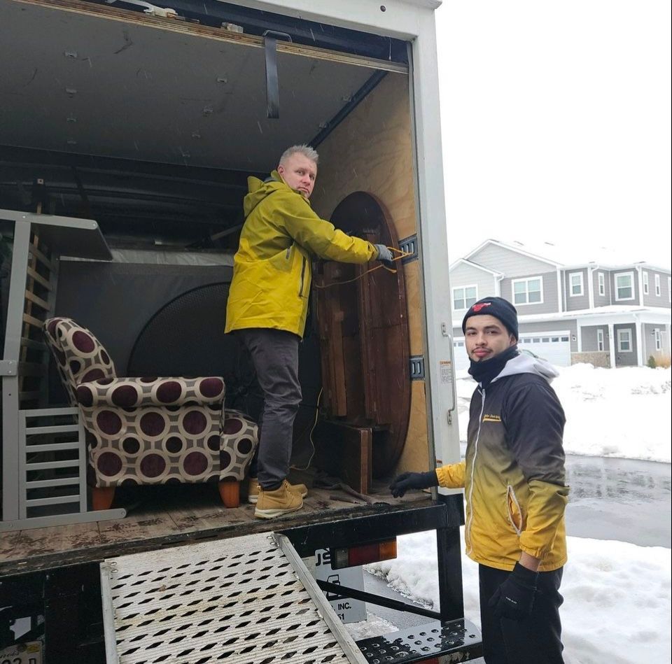 Two people loading furniture into a truck on a snowy day. One holds a table, the other stands next to a ramp.