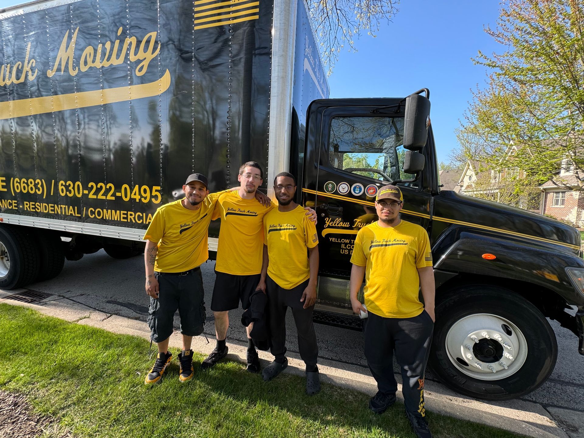 Four movers in yellow shirts stand in front of a black moving truck.