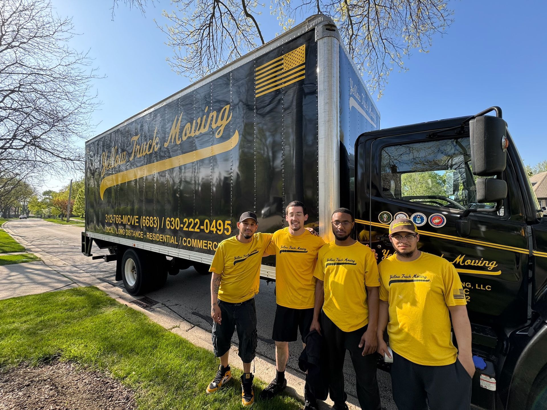 Four movers in yellow shirts stand in front of a black moving truck on a sunny day.