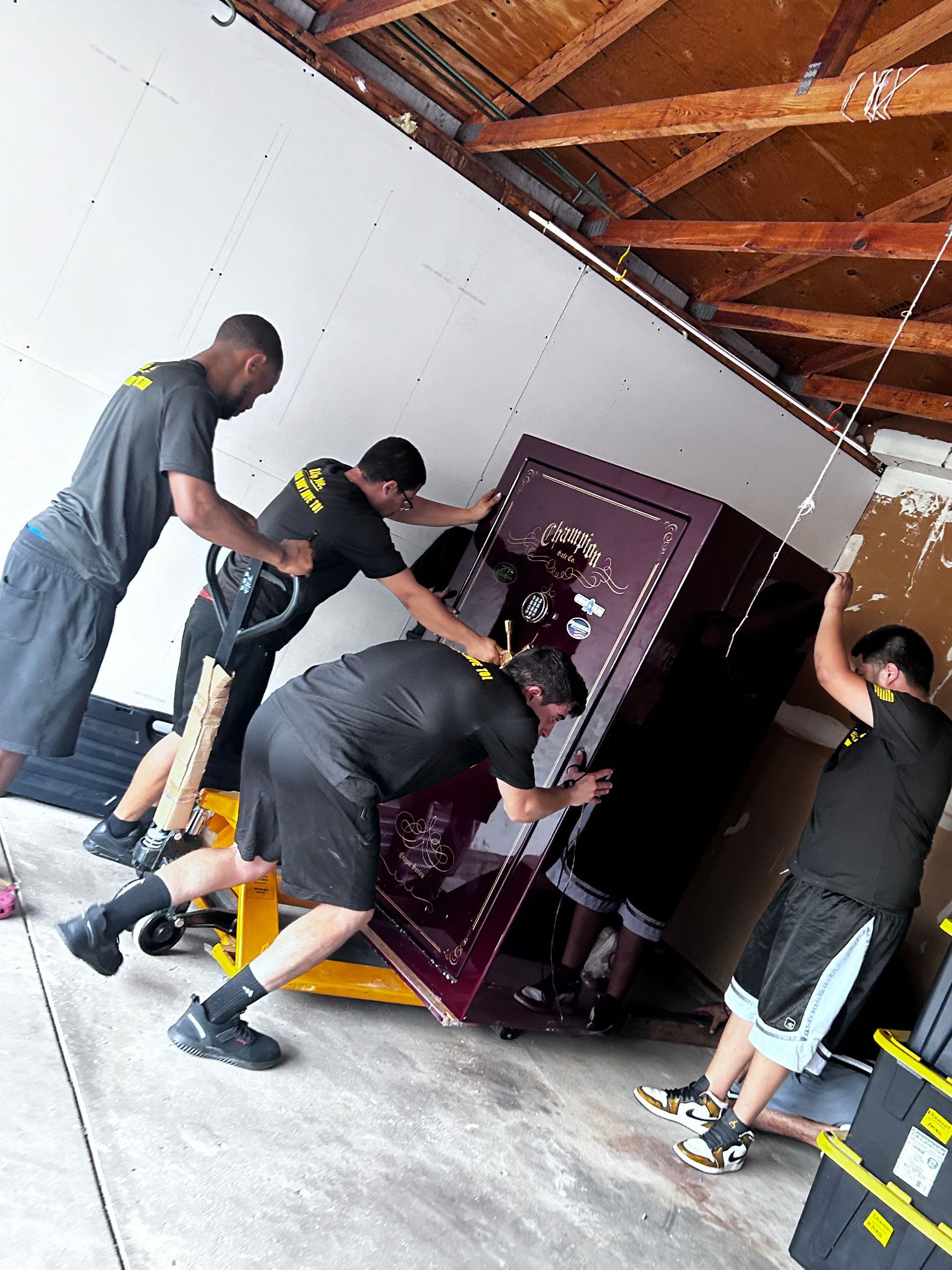 Four movers using a pallet jack and pushing a large, burgundy safe through a garage.