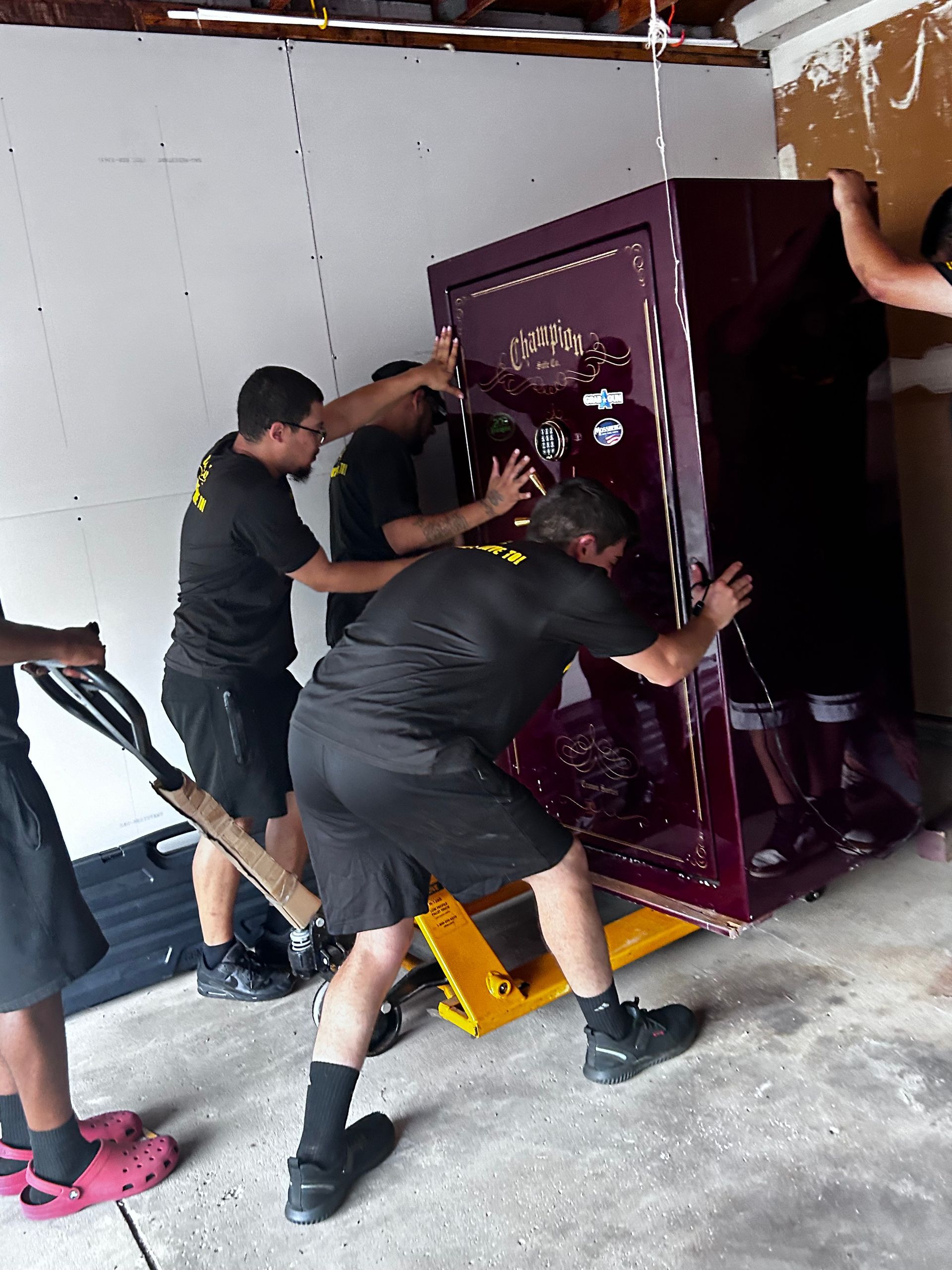 Four people in black shirts are pushing a large purple safe on a yellow dolly. They are in a garage.