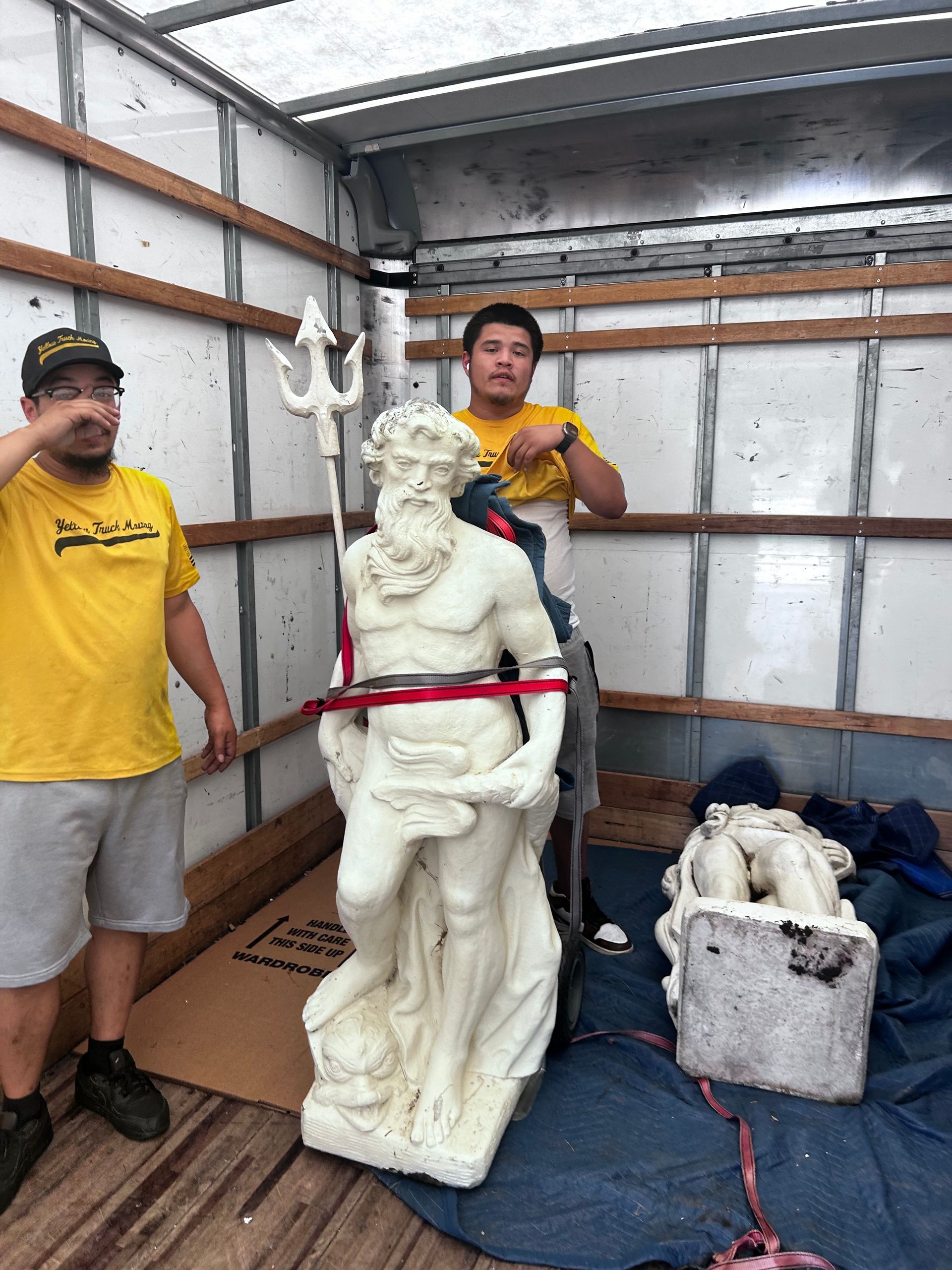 Two men transporting a white statue of a man holding a trident in a truck. One man gestures, other watches.