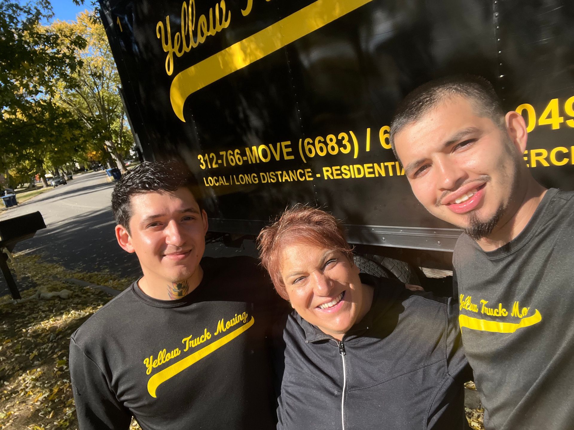 Three people smiling in front of a Yellow Truck Masters truck. Two men wear black shirts, one with a woman in between them.