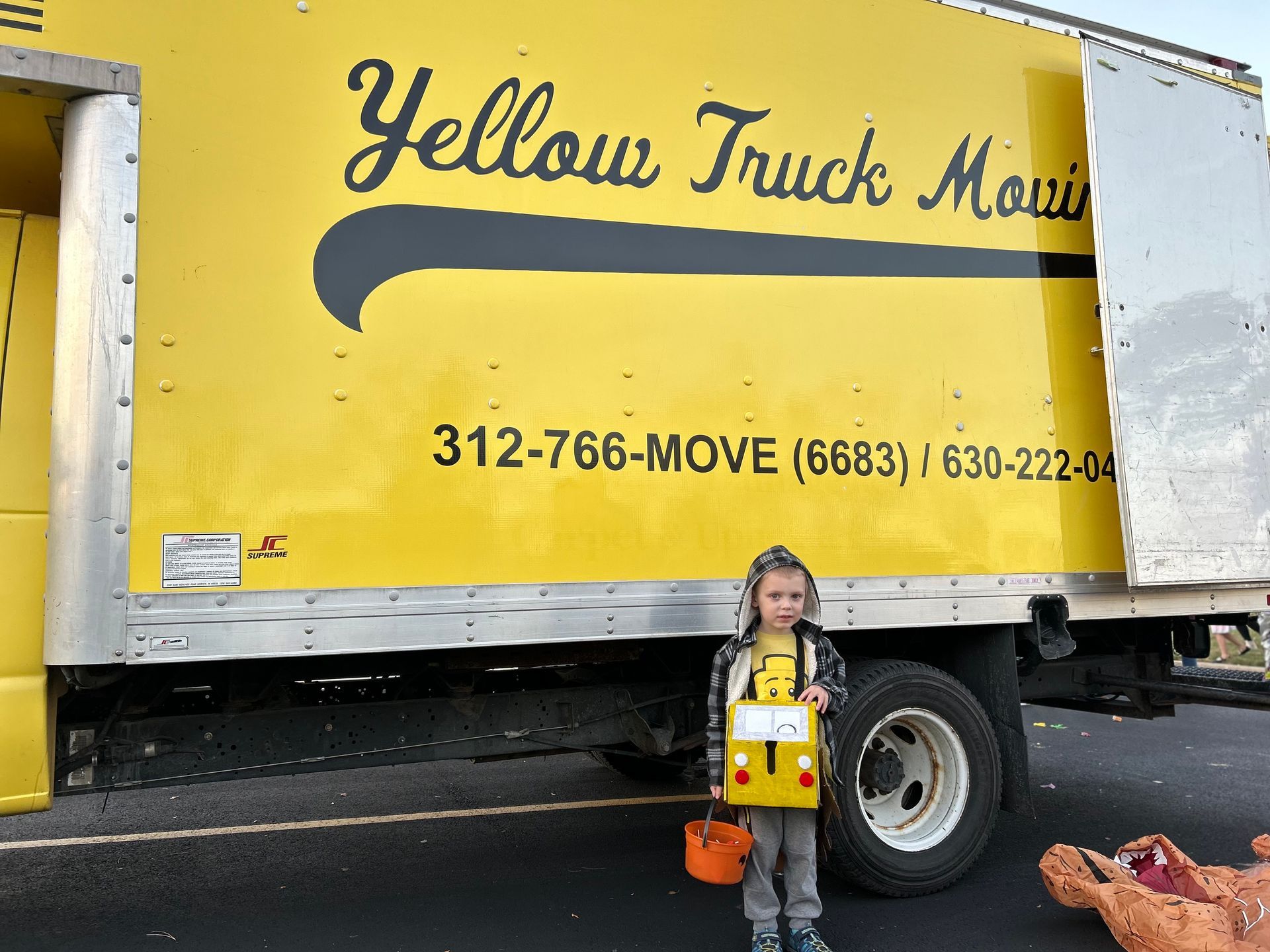 Child in a truck costume stands in front of a Yellow Truck Moving truck.