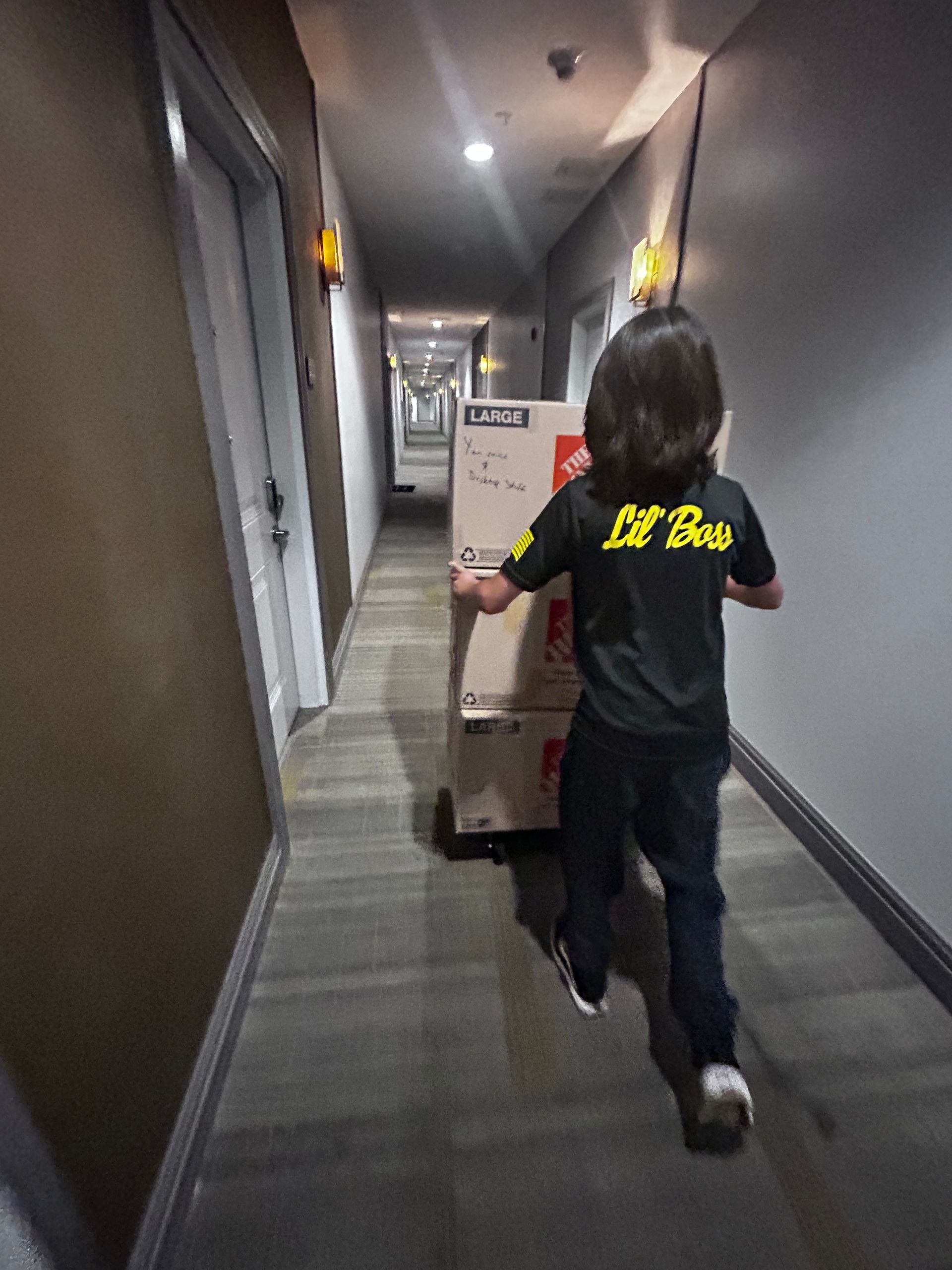 A person, wearing a black shirt, pushing two large boxes down a hallway.