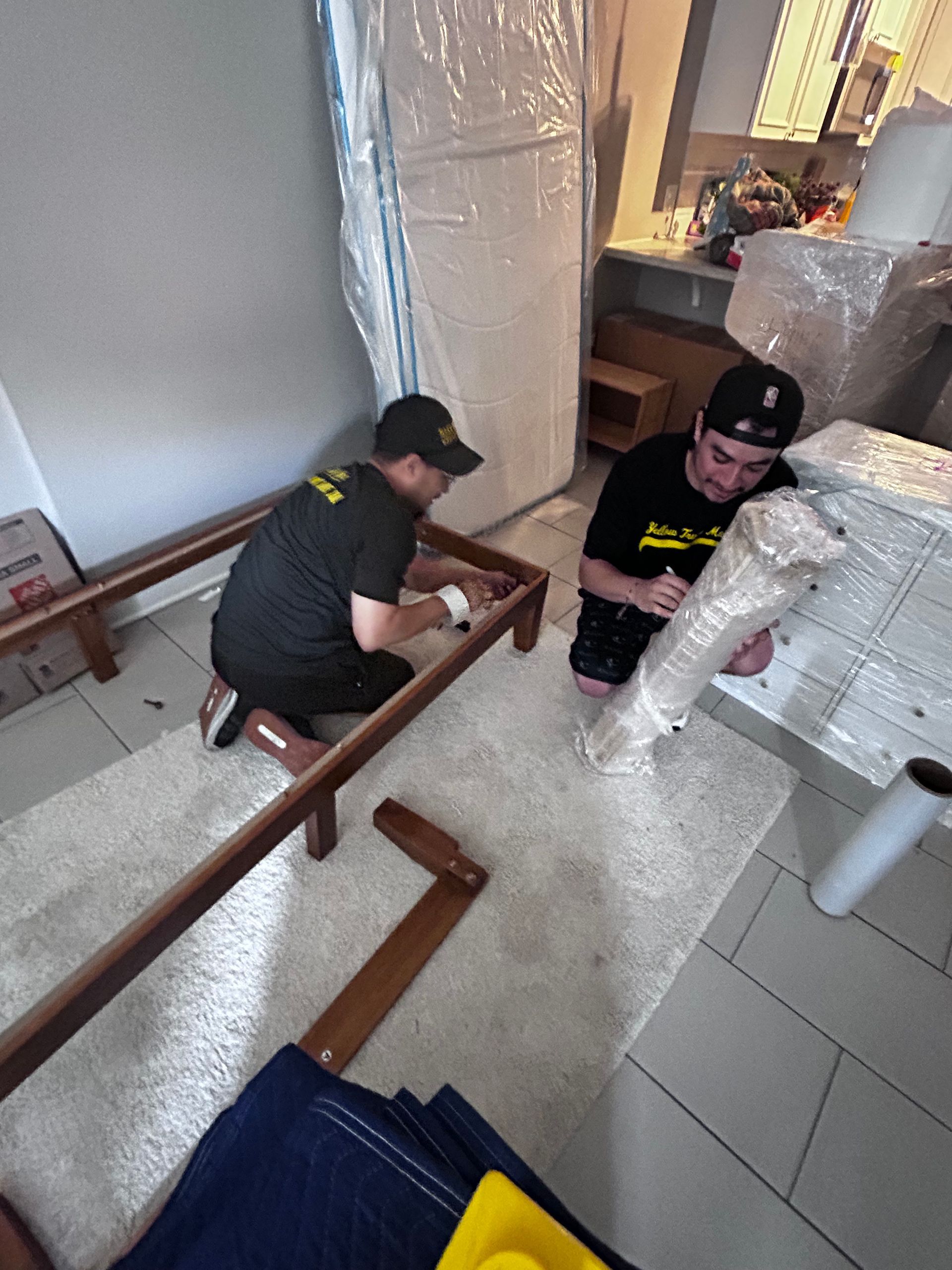 Two movers in black shirts assembling a bed frame on a white rug. Furniture wrapped in plastic.