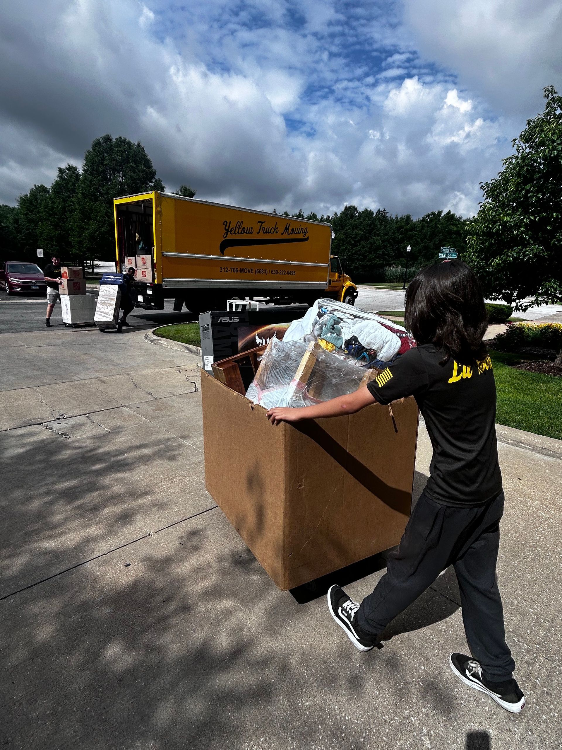 Person pushing a cardboard box filled with items towards a yellow moving truck on a sunny day.