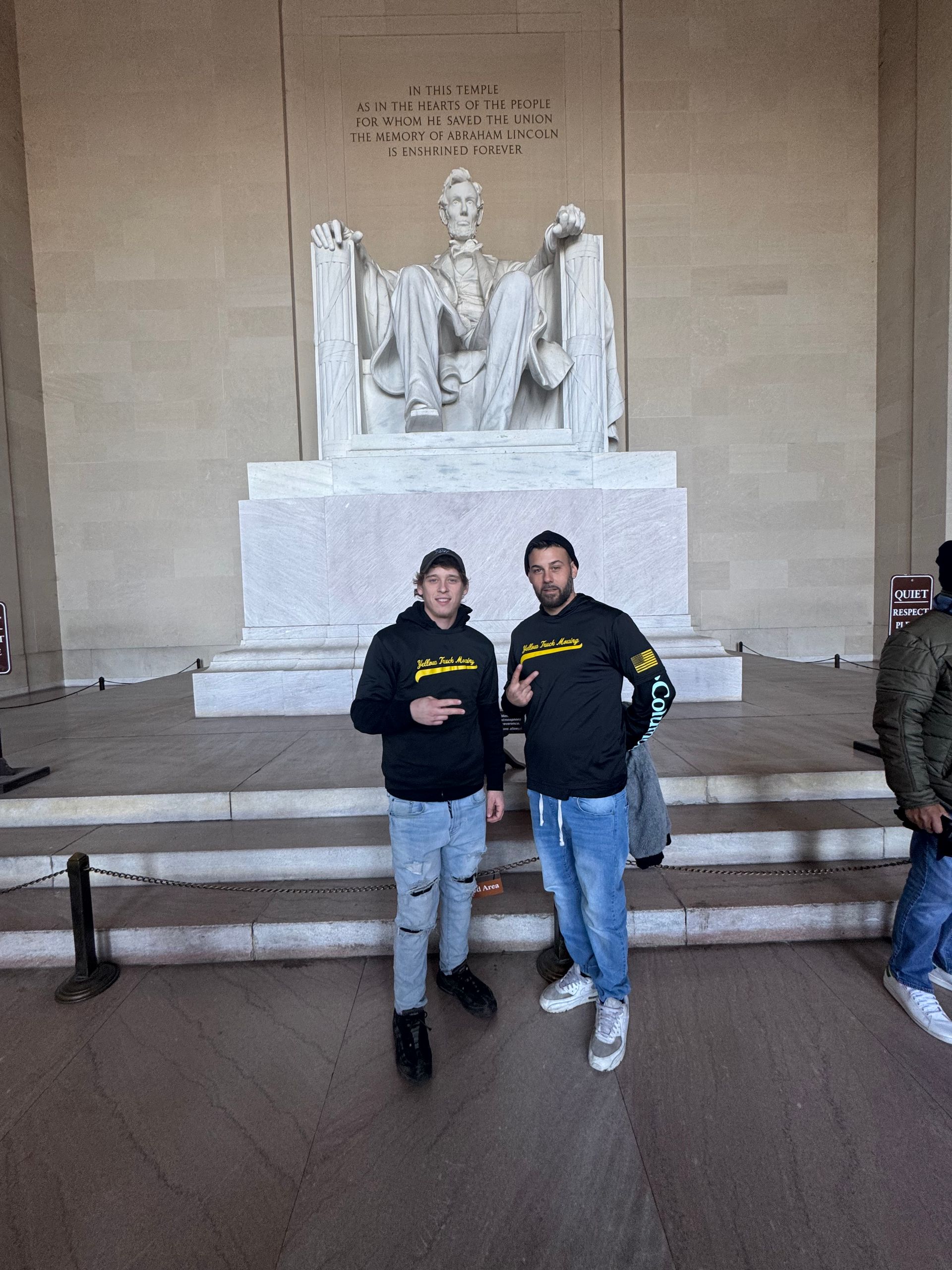 Two men pose in front of the Lincoln Memorial statue. Both wear hoodies; one makes a peace sign.