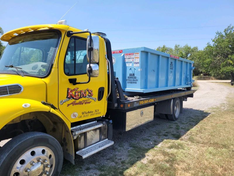 A yellow tow truck is carrying a blue dumpster.