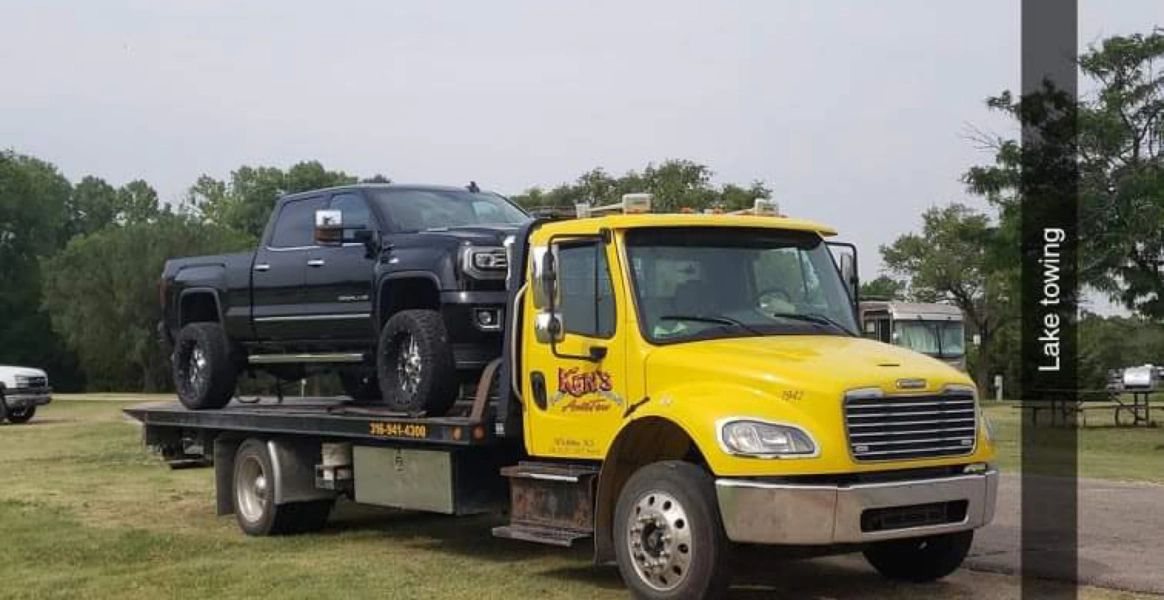 A yellow tow truck is carrying a black truck on its flatbed.