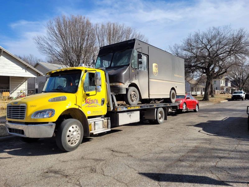 A yellow tow truck is towing a delivery truck down a street.