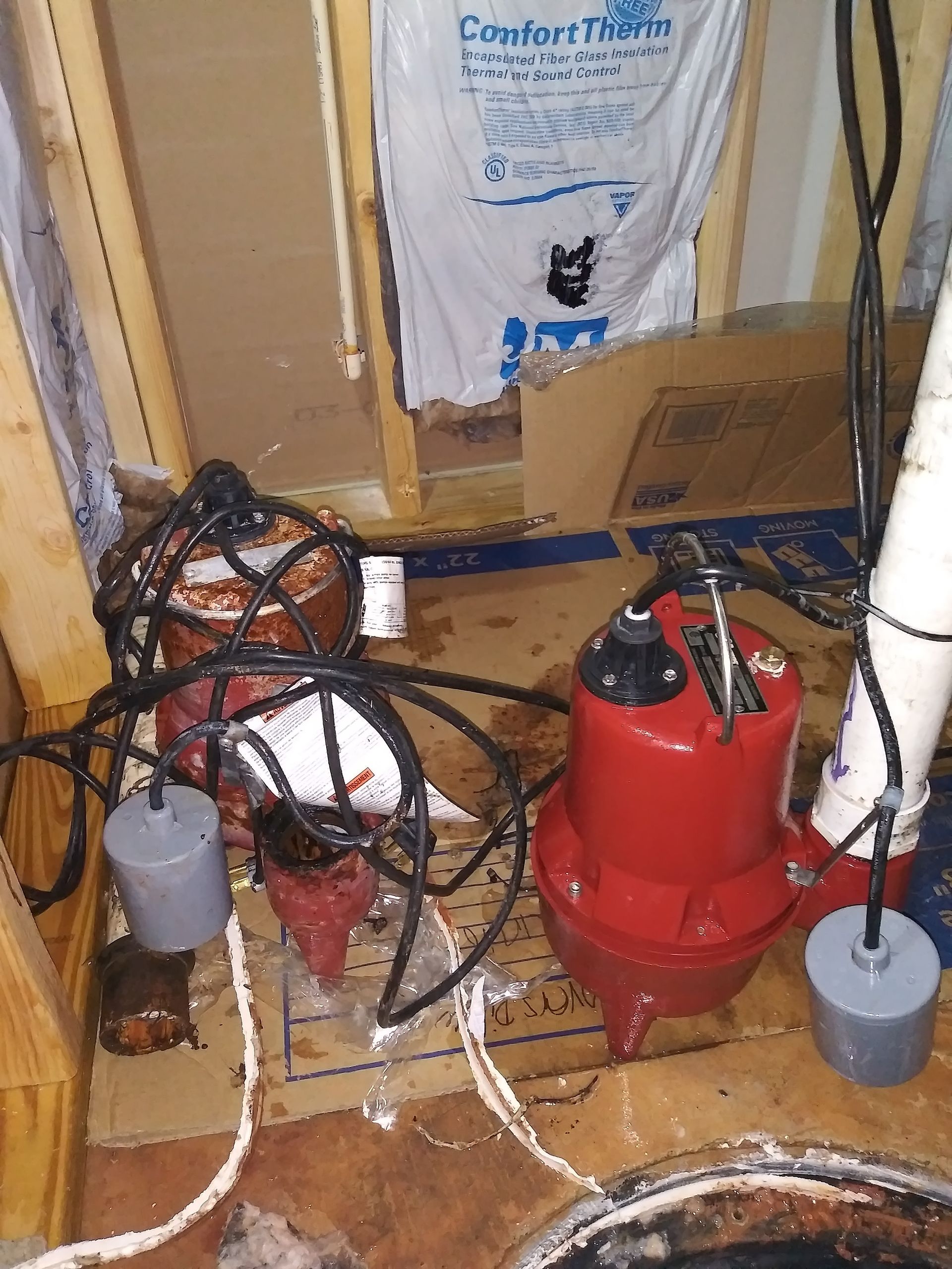 Two red sump pumps with cords and floats in a basement, next to insulation.