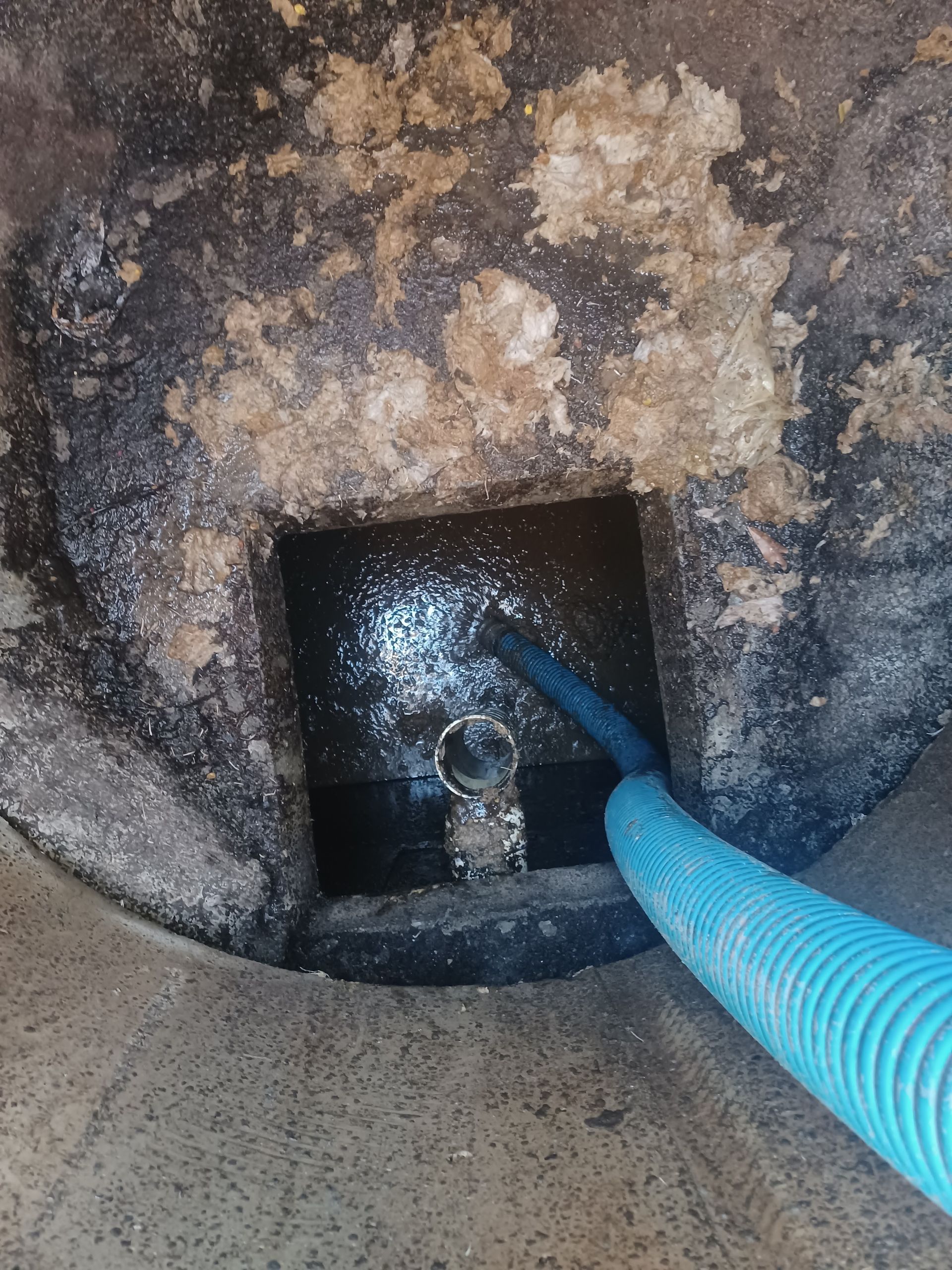 Blue hose entering a dark square pit, surrounded by dirt and white particles, inside a larger concrete cylinder.