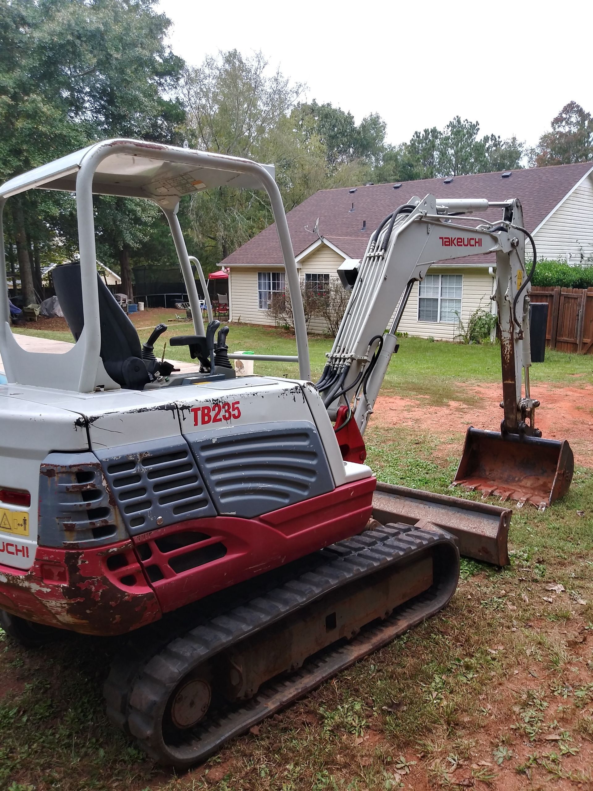 A small, white and red excavator with tracks parked on grass, in front of houses and trees.
