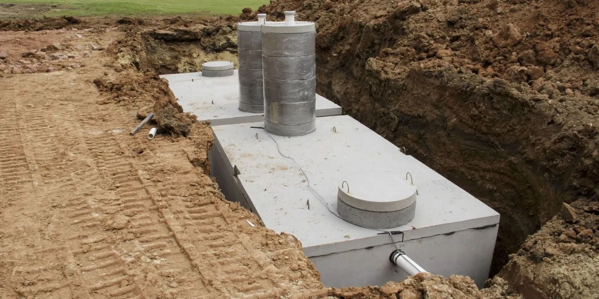 Concrete septic tank installed in a dug trench. Two vents and access hatches visible. Earth surrounds.