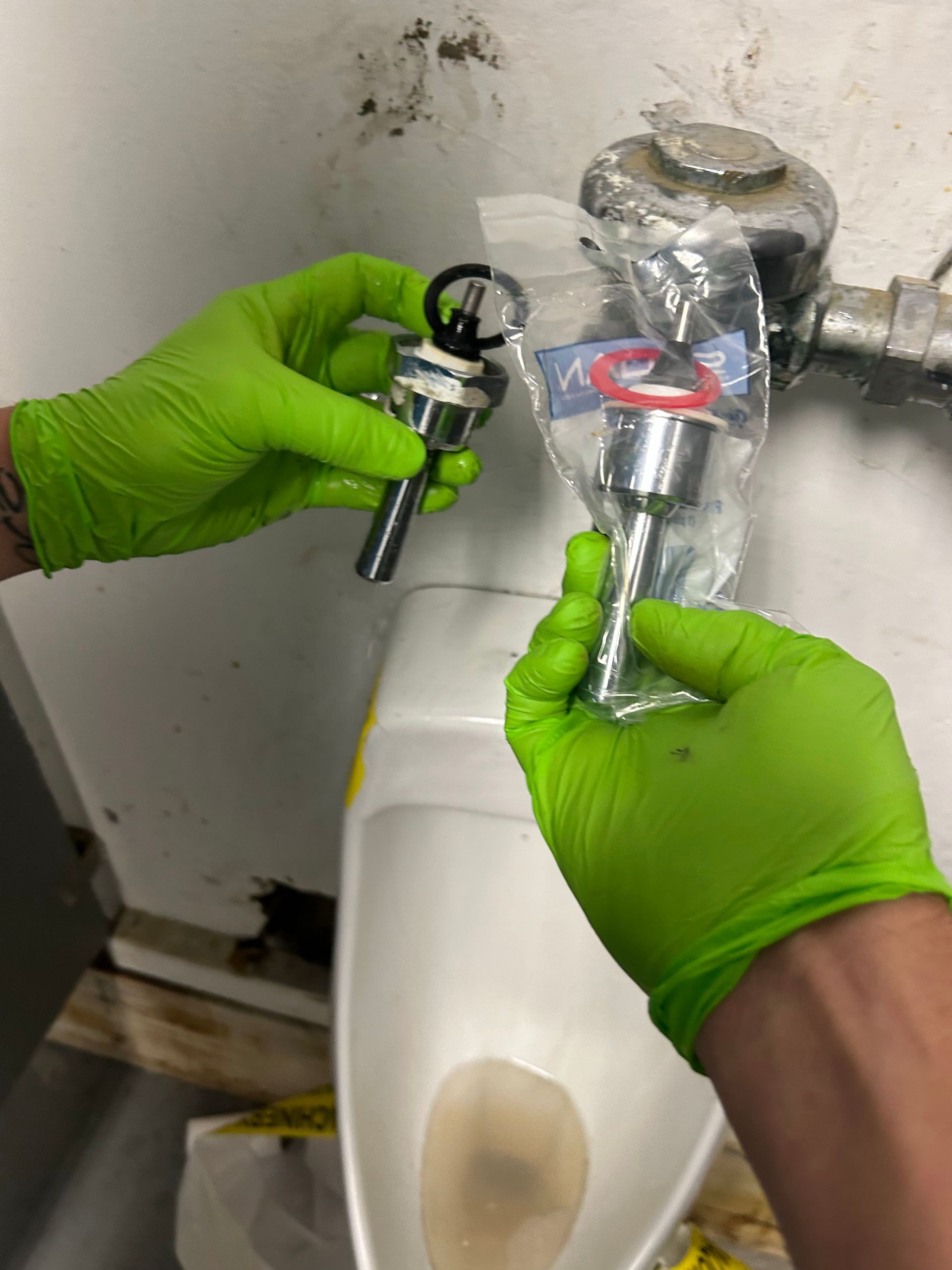 Hands in green gloves holding two urinal flush valves near a urinal.