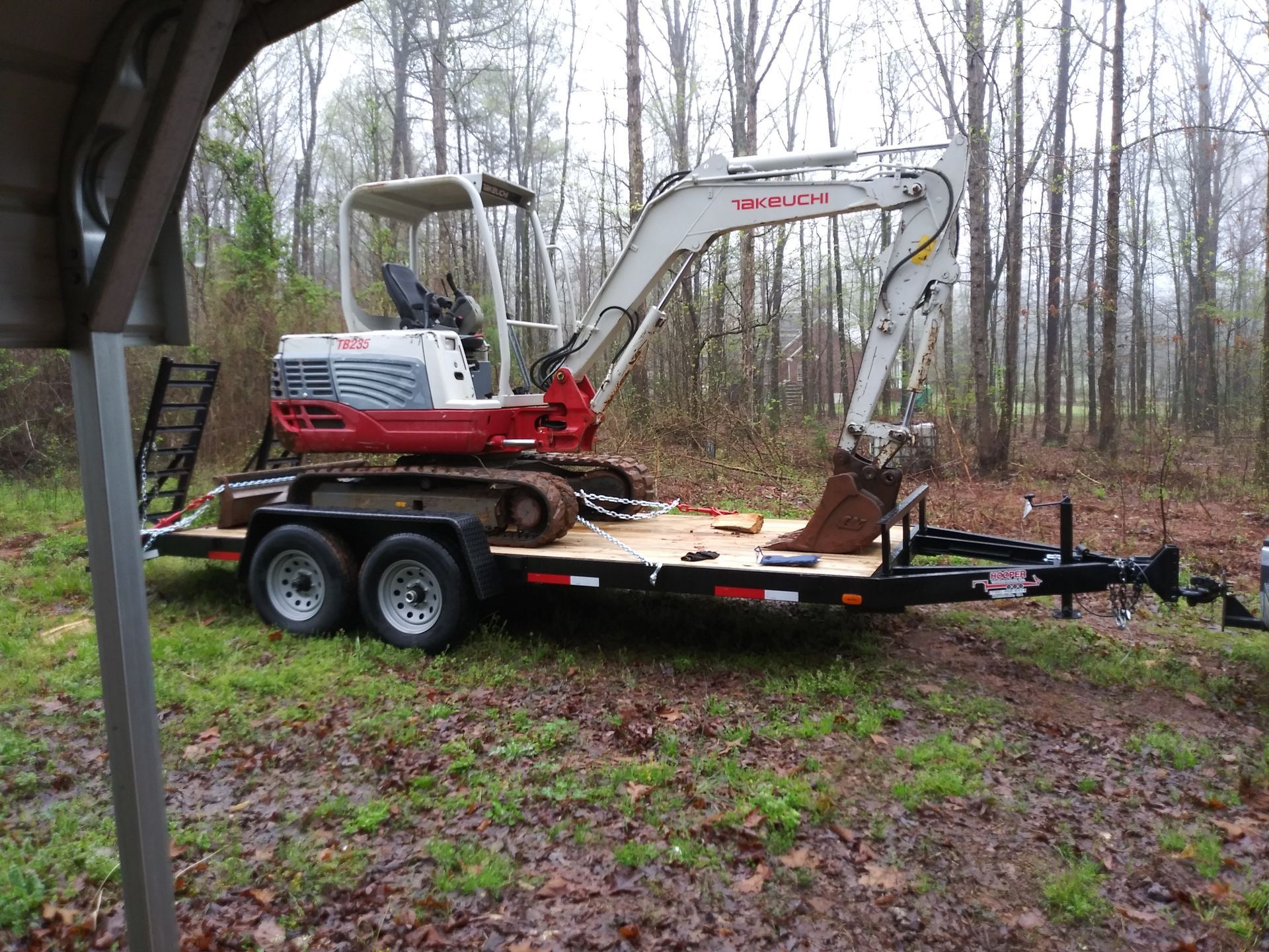 Mini excavator on a trailer parked in a wooded area. Red and white machine, black trailer, overcast day.