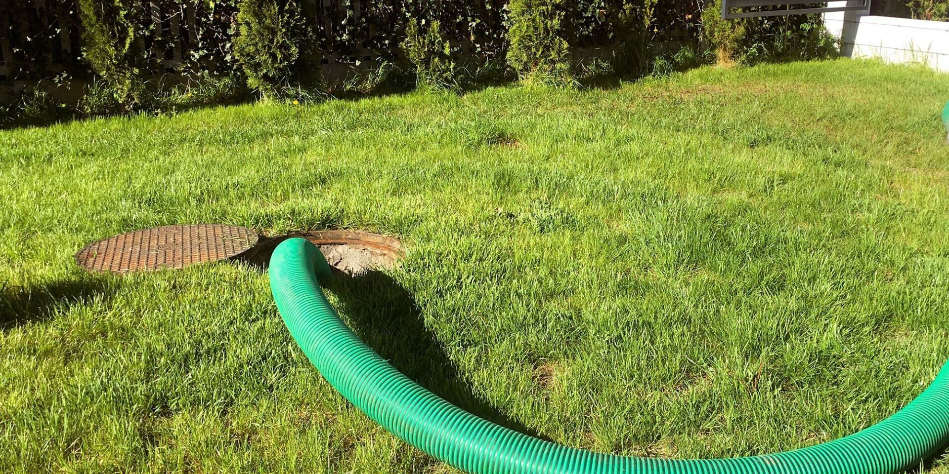 Green hose connected to an open, circular lid in a grassy yard.