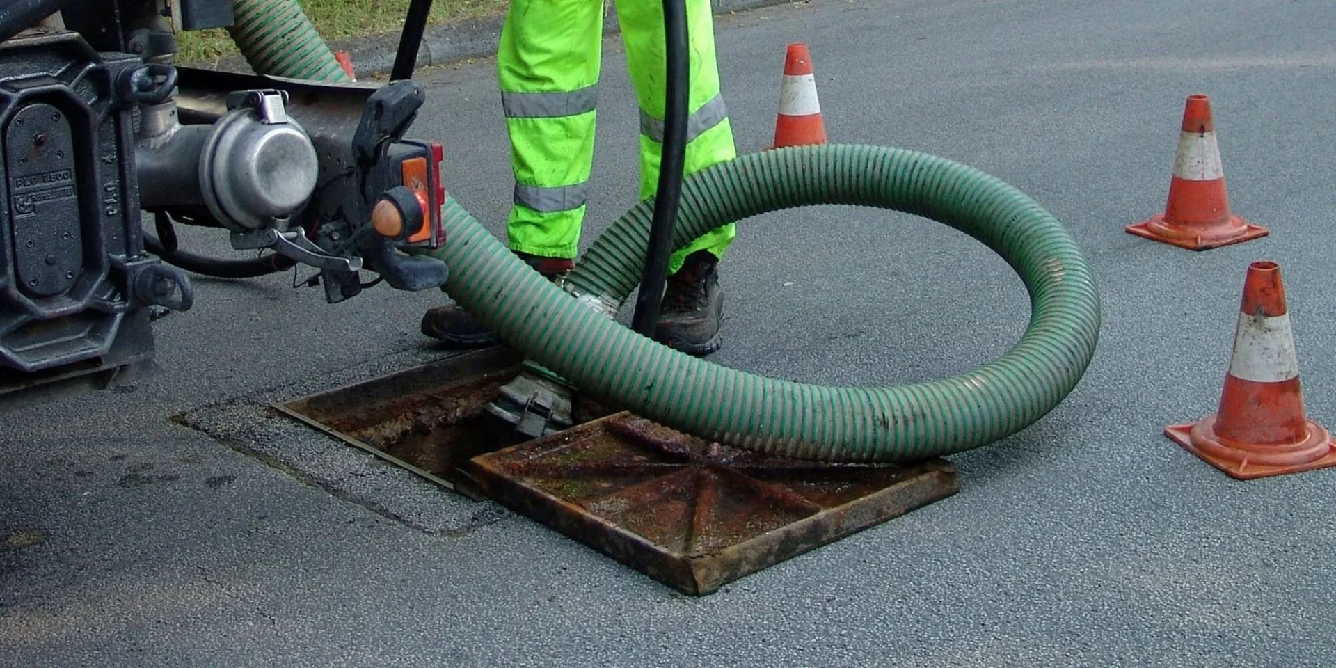 A worker in neon green overalls uses a hose to clean a manhole in the asphalt road. Cones mark the work area.