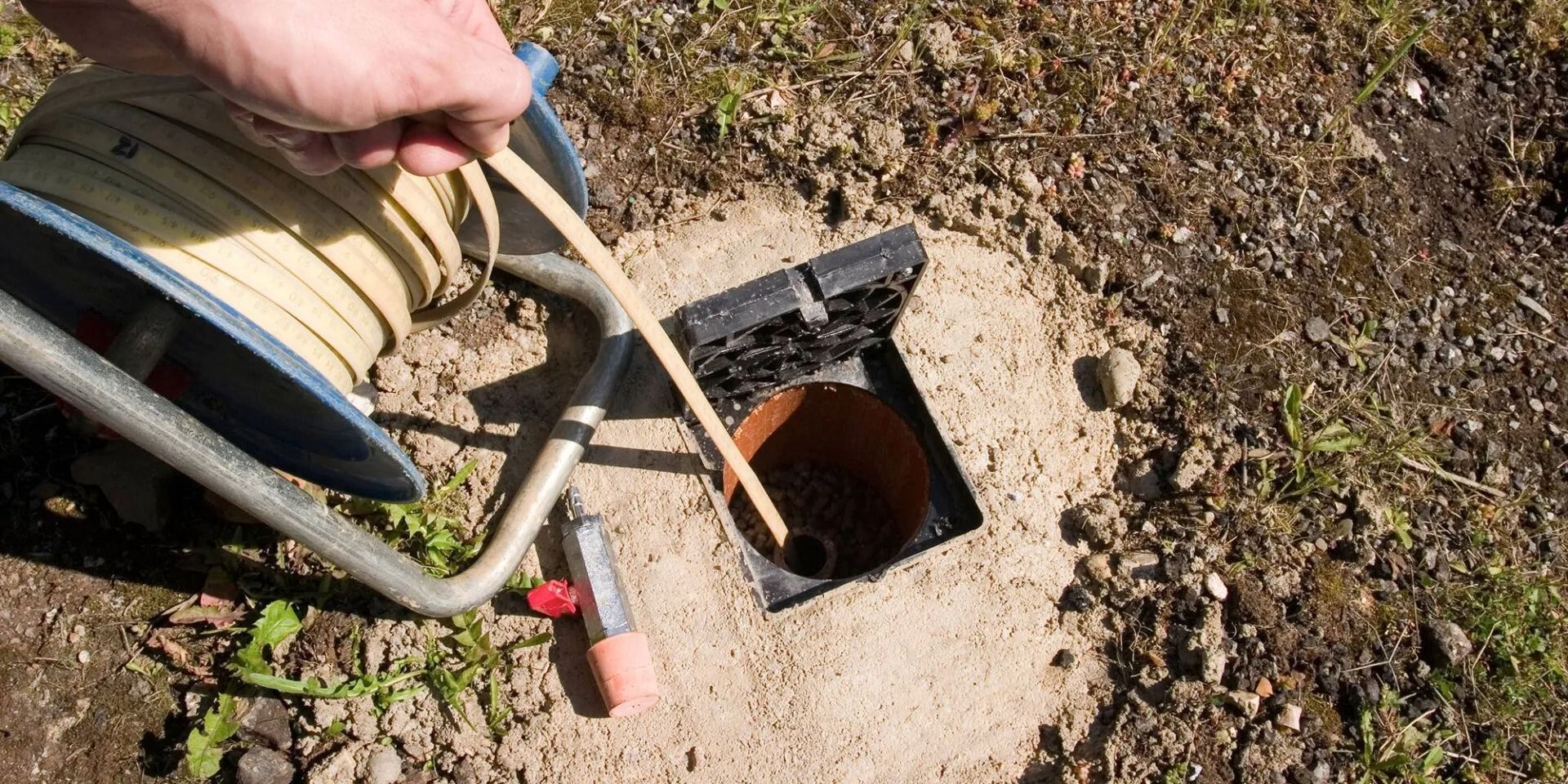 A person uses a stick to check an open drain in the ground near a hose reel.