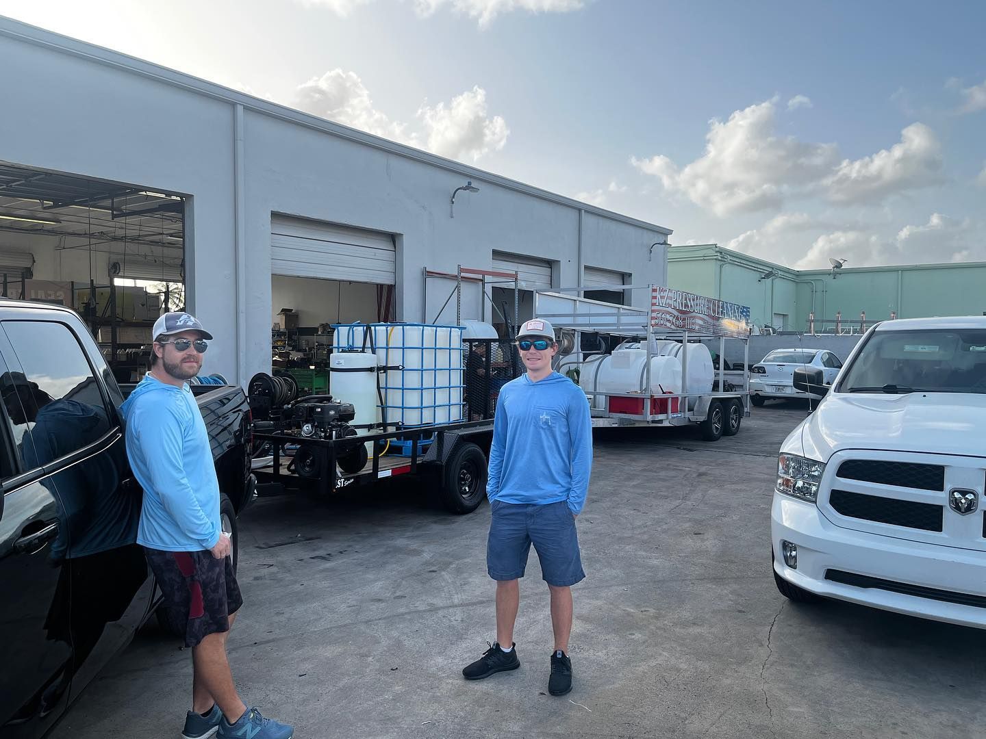Two men standing in an industrial lot beside trucks and a warehouse under a cloudy sky