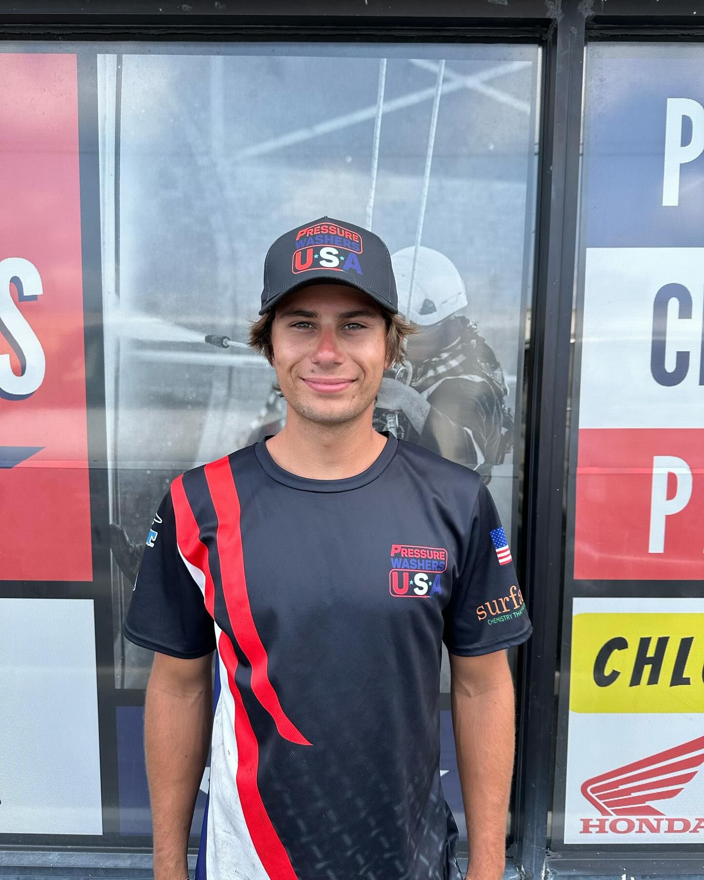 Smiling man in black and red team shirt and cap standing in front of a glass storefront.
