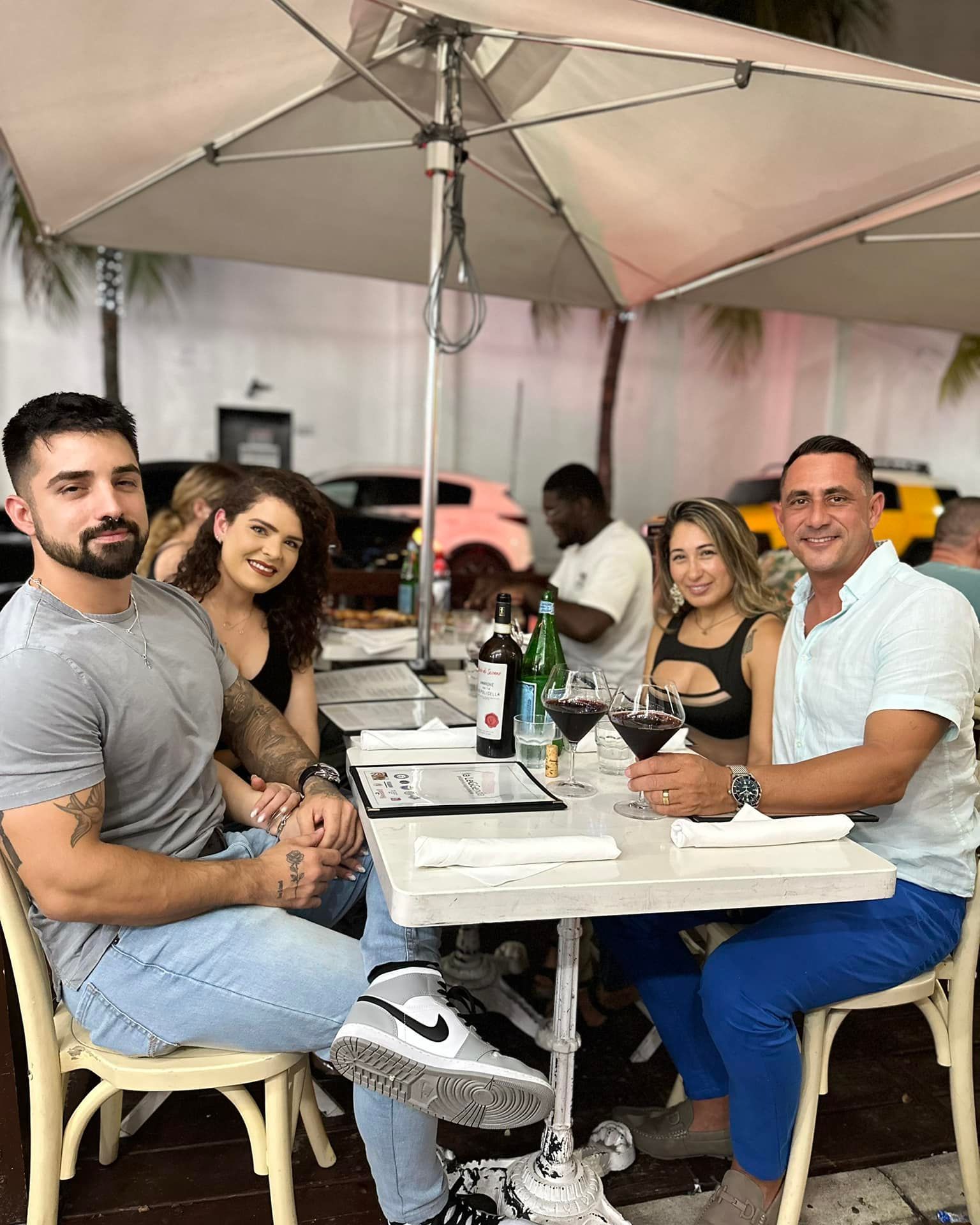 People seated at an outdoor café table under a large umbrella, smiling and socializing.