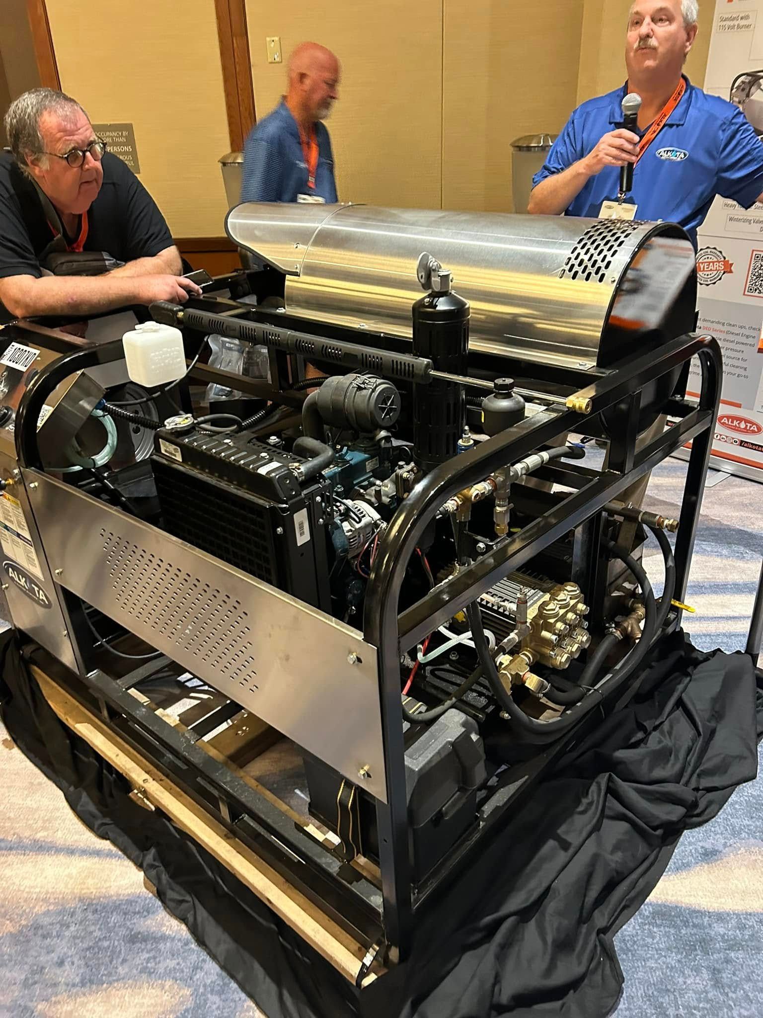 Large black machinery with a gold cylinder on display at a conference, with people standing nearby