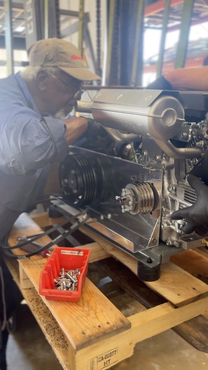 Worker inspecting machinery on a wooden pallet, with tools and metal components in a workshop.