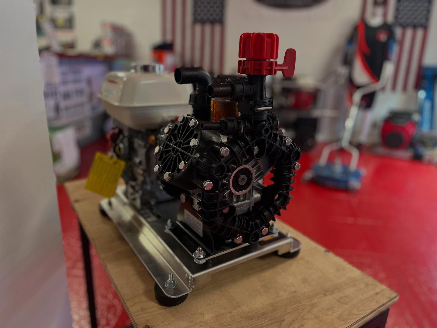 Small model engine mounted on a wooden base, with a red valve assembly in a workshop setting.