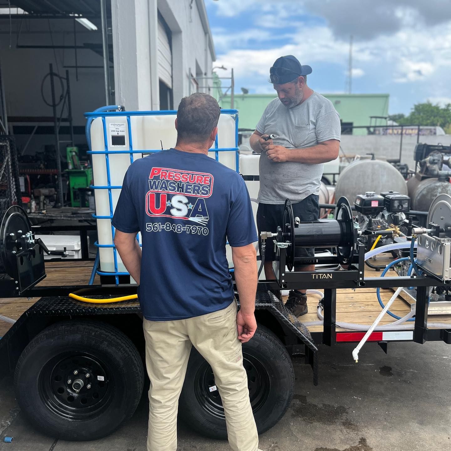 Two men work on machinery at a trailer-mounted workbench outside a shop.