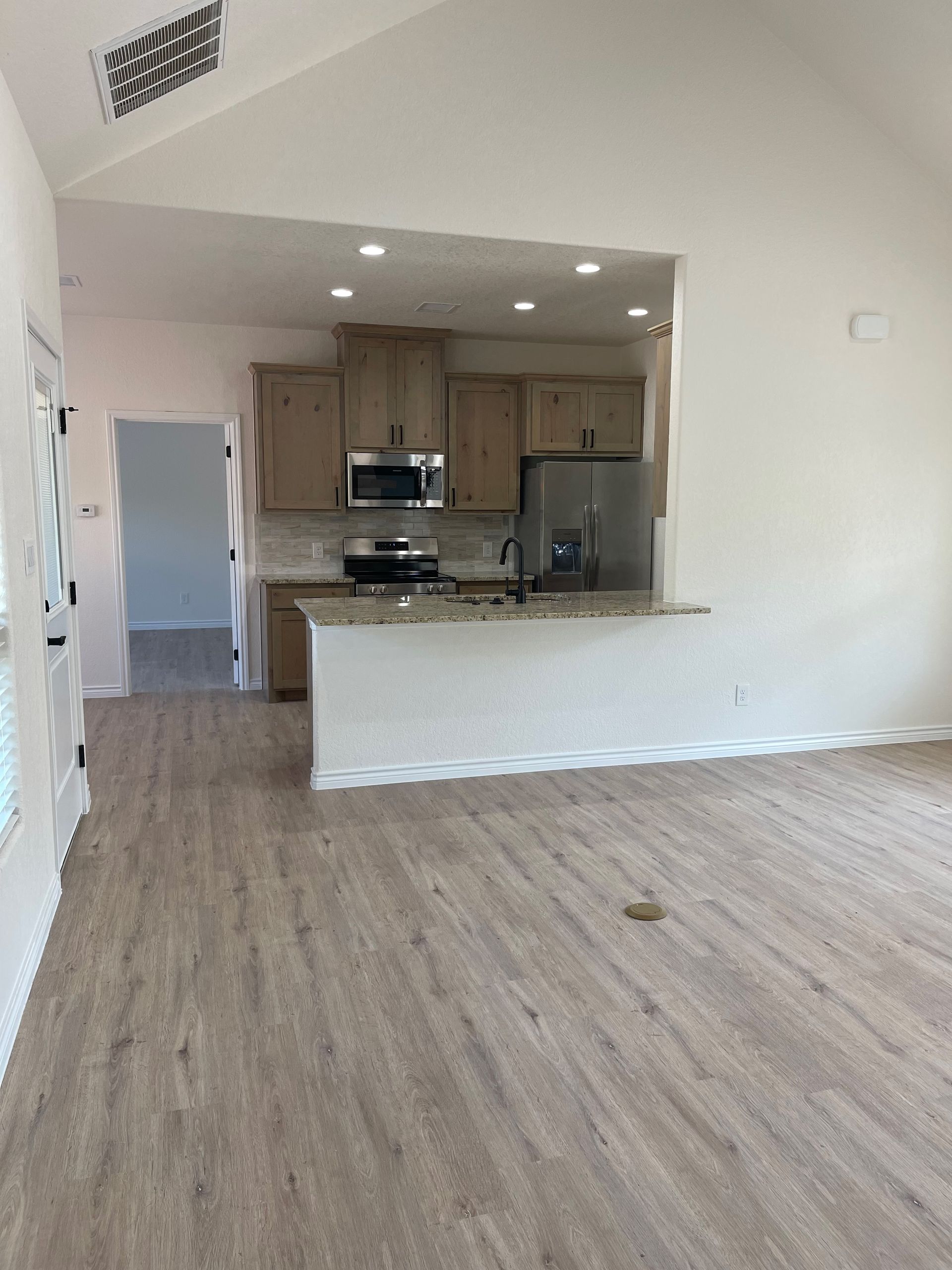 A large empty room with hardwood floors and a kitchen in the background.