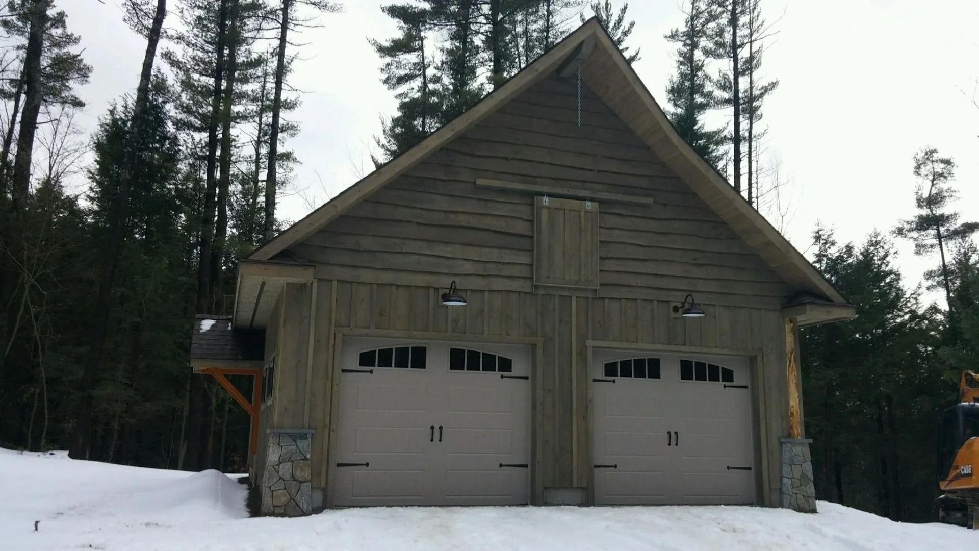 Wooden two-car garage with snow on the ground, surrounded by tall trees