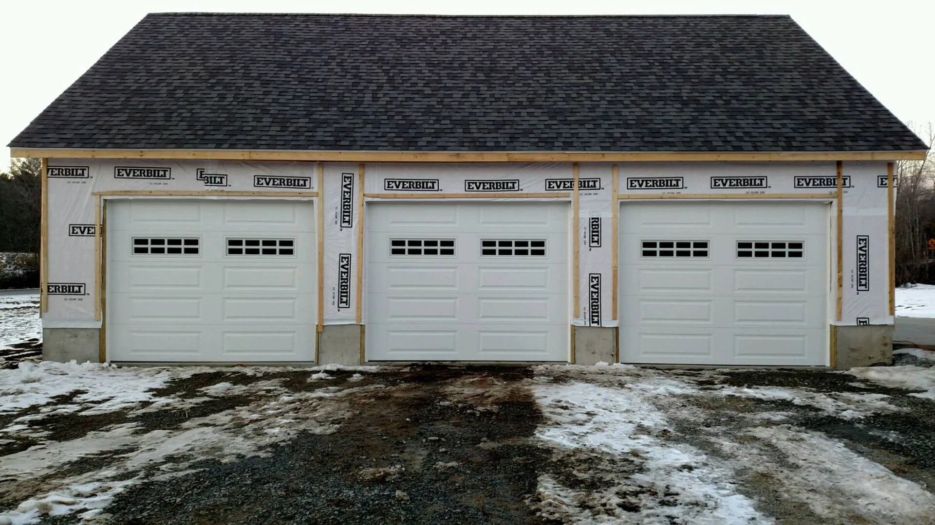 Three white garage doors on a beige building with a dark shingled roof and light snow on the ground