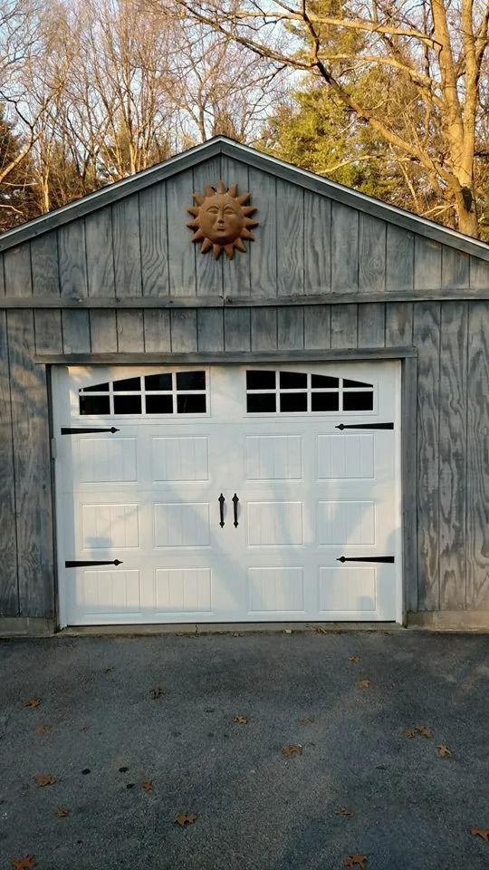 White double garage door on a gray barn-like outbuilding with a sun decoration above, surrounded by trees.