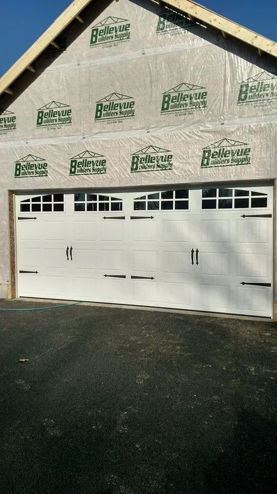 White garage door on a framed house under construction, wrapped in housewrap, with a dark asphalt driveway.