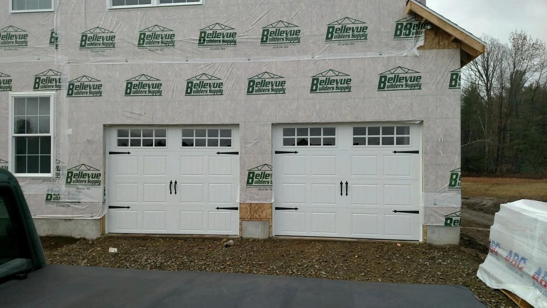 Two white garage doors on a house under construction with unfinished siding and stone veneer below.