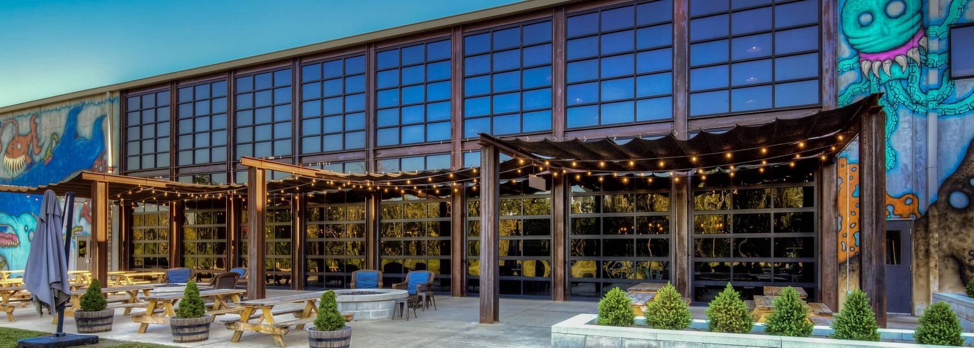 Modern glass-front restaurant with a wooden pergola, potted plants, and colorful mural art outdoors