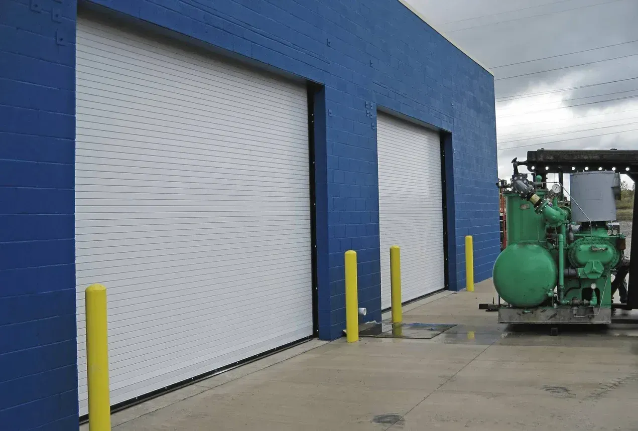 Blue industrial building with white garage doors, yellow bollards, and green machinery outside on a wet concrete lot
