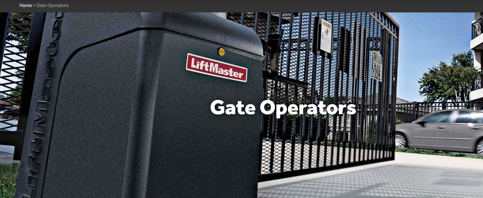 Gate Operators sign on a black metal gate by a driveway, with cars and buildings in the background.