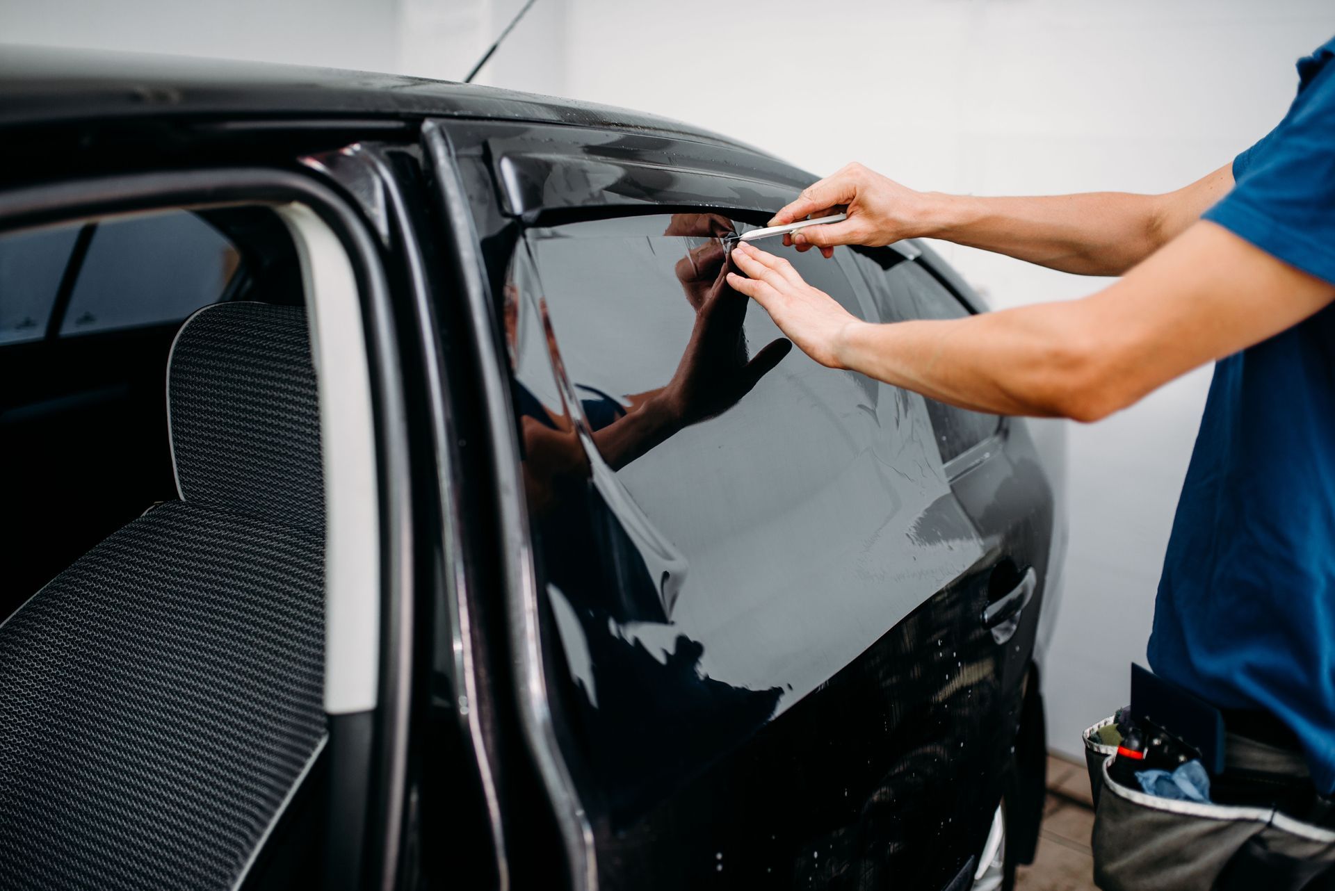 A technician in a blue shirt applies a dark, protective film to a car’s side window.