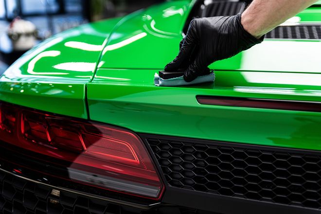 A gloved hand uses a foam block to apply a protective coating to the glossy green rear deck of a sports car.