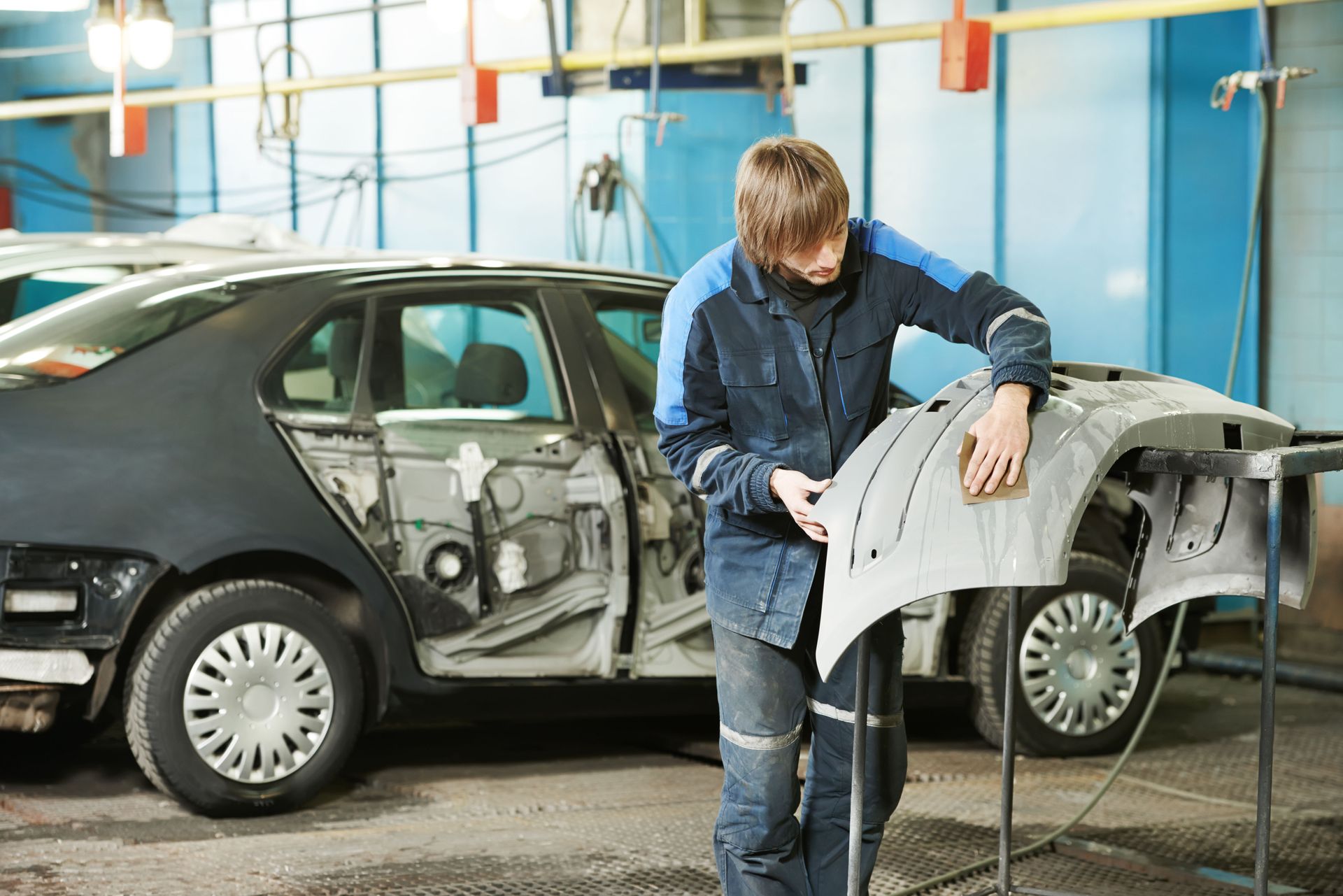 A worker in blue workwear sanding a detached car bumper in an automotive repair shop with a car body in the background.