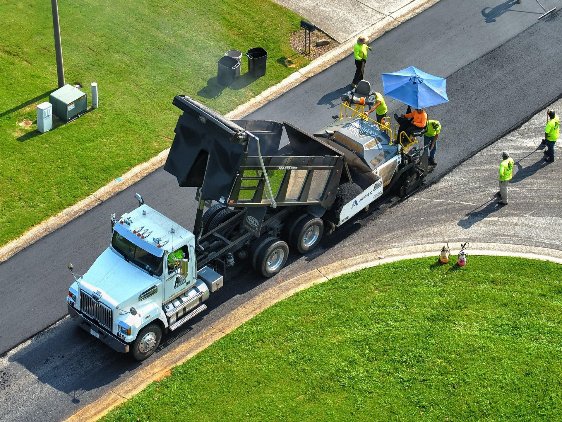 Asphalt paving in progress: Truck dumping asphalt into a paving machine on a road. Workers in vests.