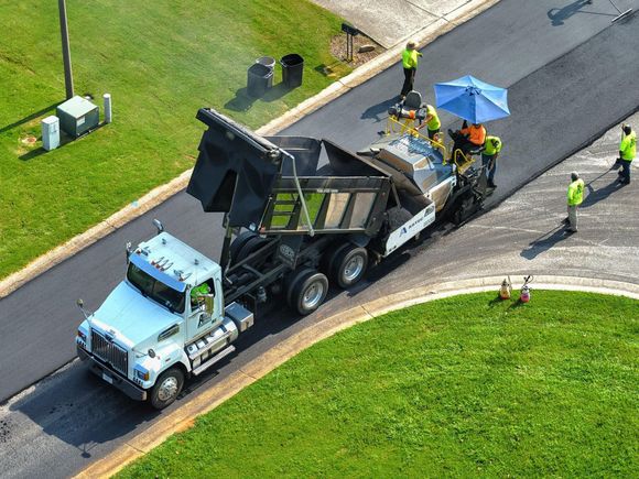 Asphalt paving in progress: Truck dumping asphalt into a paving machine on a road. Workers in vests.