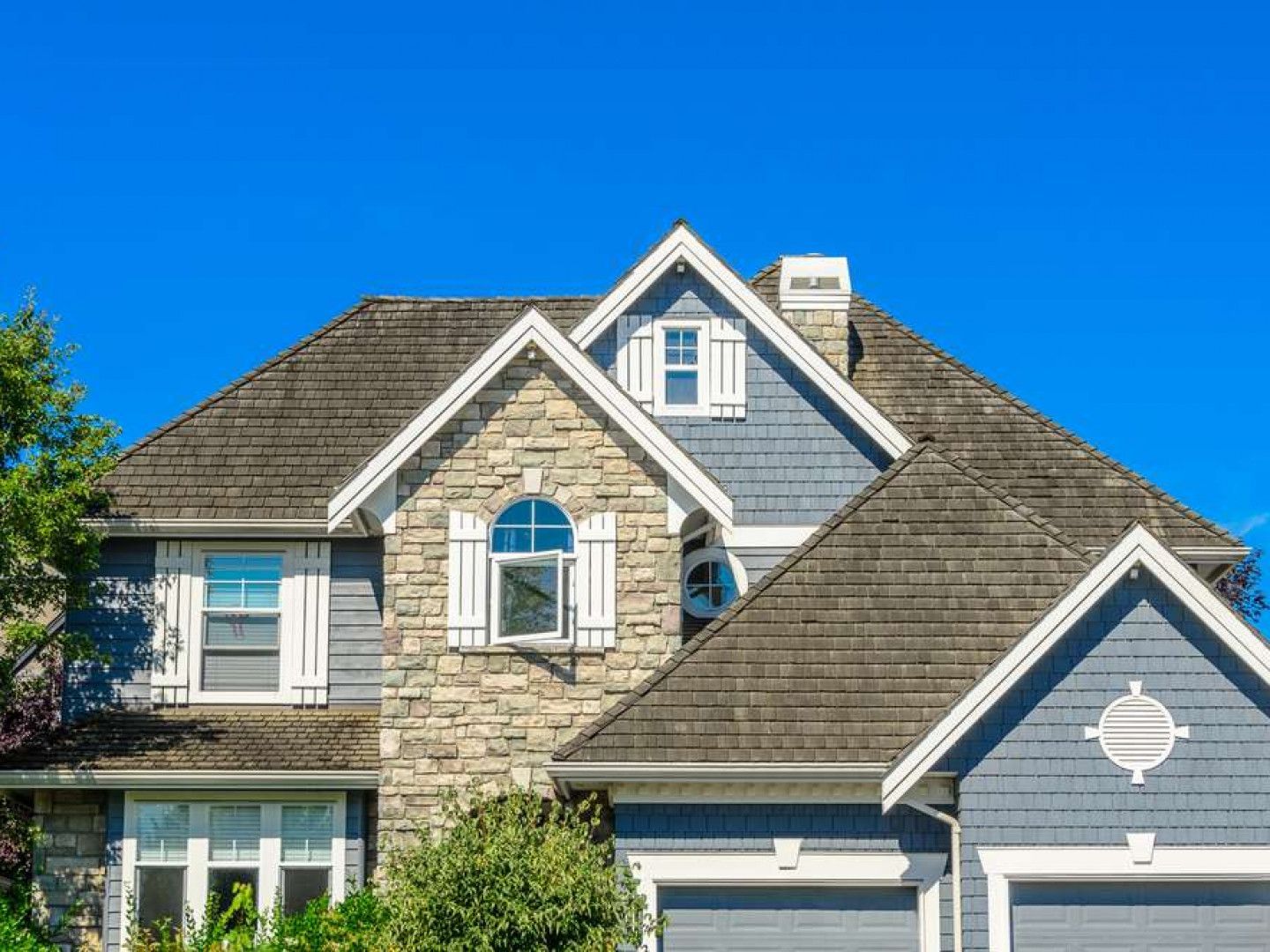 A two-story suburban house with stone veneer, blue shingle siding, a dark roof, and a garage under a clear blue sky.