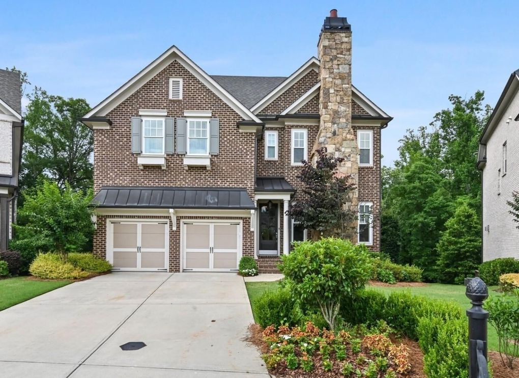 Two-story brick house with a stone chimney, two-car garage, and landscaped front yard under a blue sky.