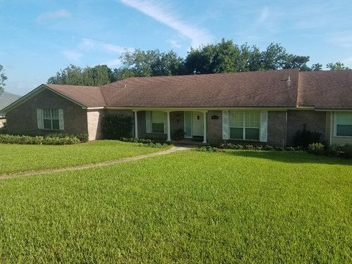 A single-story, tan brick house with a dark brown shingled roof and large windows behind a wide, grassy front lawn.